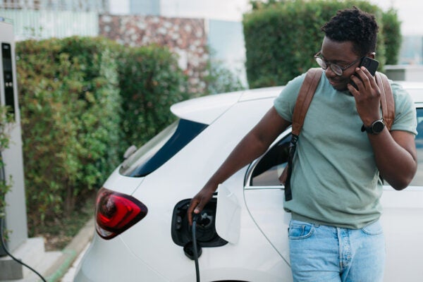 Handsome young adult man standing next to his electric vehicle and using his smart phone while his car is plugged into the charging station